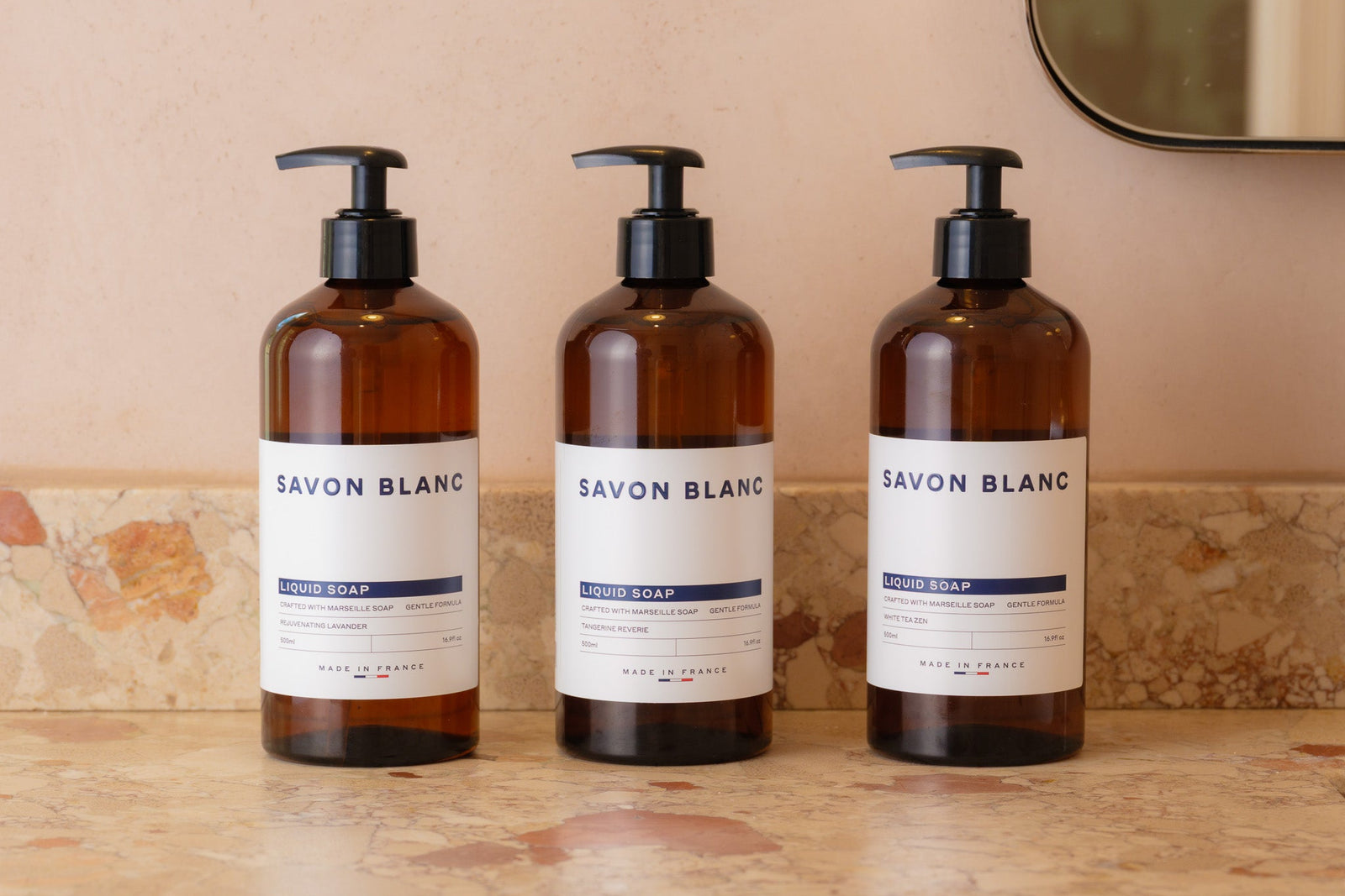 Three bottles of SAVON BLANC liquid hand soap on a wooden surface with a beige wall in the background.